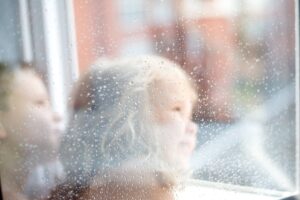 Two children looking at rain, which has delayed their parent's summer house paint project.