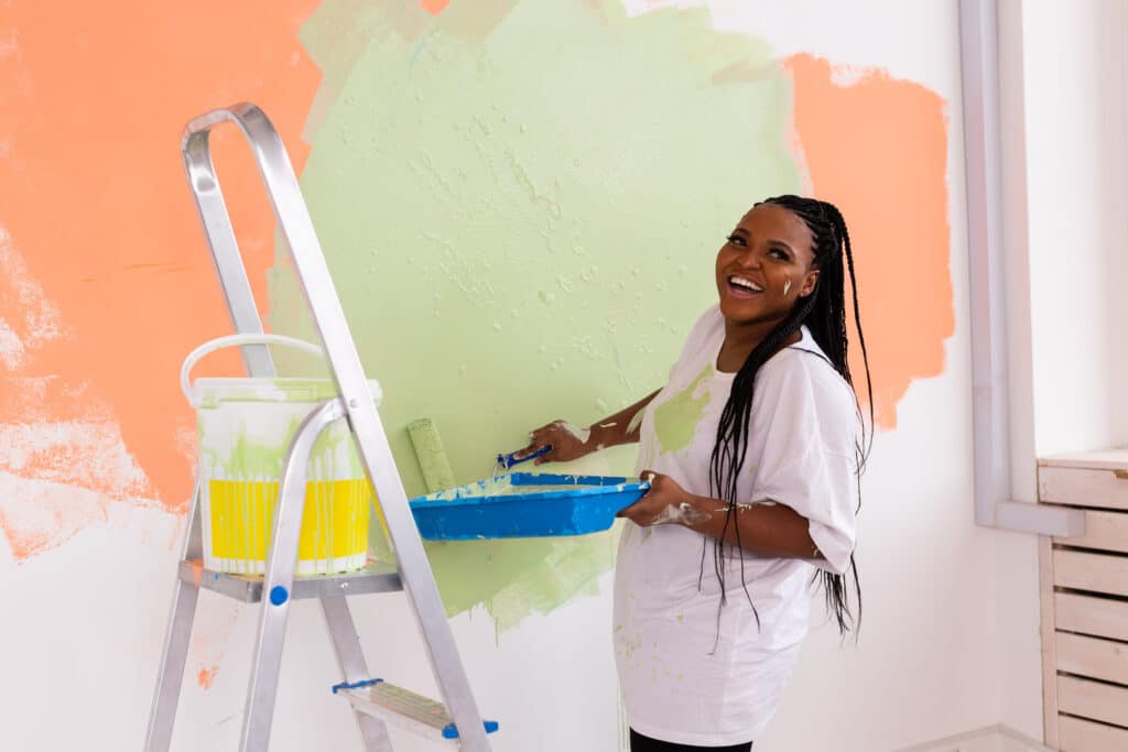 A joyful woman with a wide smile painting a wall in a soothing light green shade, symbolizing QUALITY GUARANTEES with her meticulous work.