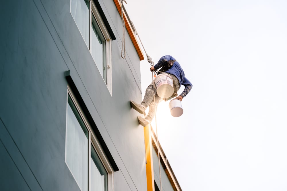 A professional painter is suspended by ropes while performing exterior commercial painting on a tall building. Wearing a safety harness and work gear, he holds paint buckets and applies fresh paint to the building’s façade. The image captures the scale and precision required for high-rise painting projects, emphasizing safety and expertise in commercial property maintenance.