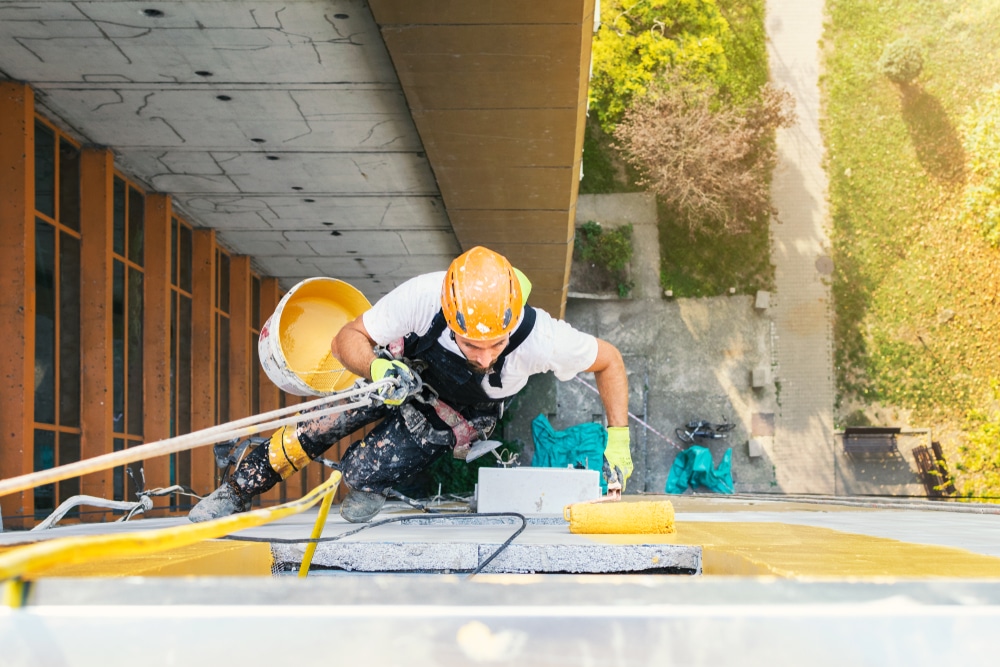 "A professional painter wearing safety gear and a harness is suspended on ropes while painting the exterior of a tall commercial building. He is using a roller brush and carrying a bucket of paint, carefully applying a fresh coat to the façade. The high-angle shot captures the worker's focus and the urban surroundings below, highlighting the precision and safety measures involved in commercial building painting projects.