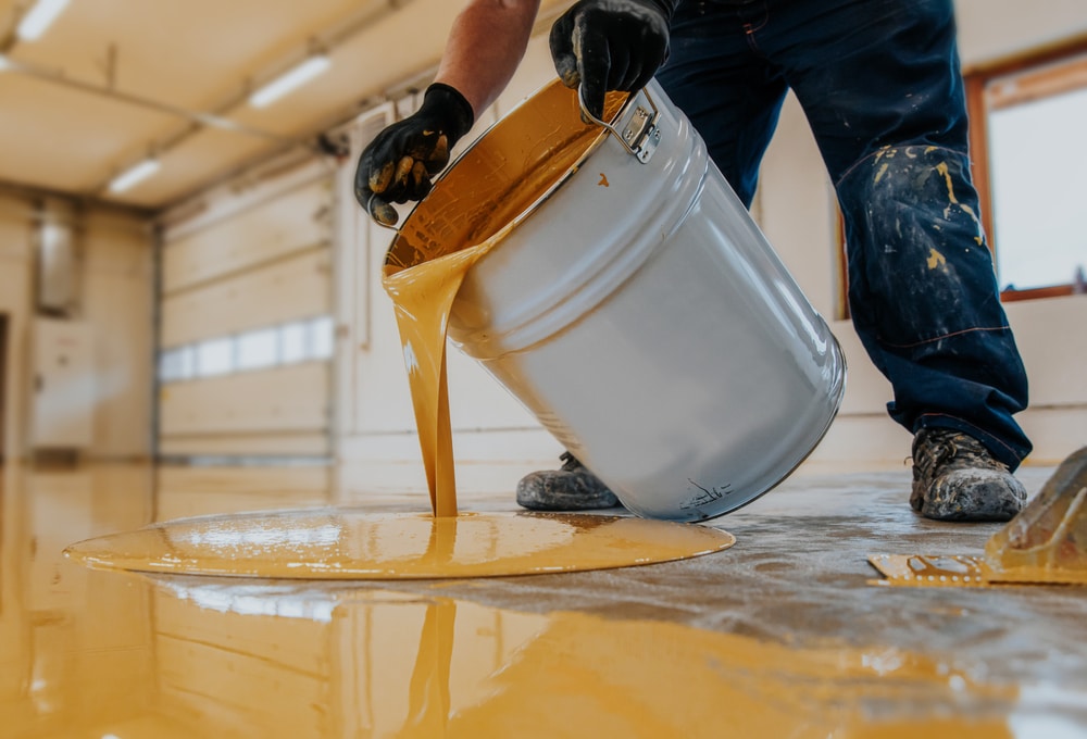 Worker pouring a large bucket of yellow industrial liquid onto a concrete floor, illustrating the application process of a protective floor coating in a commercial or warehouse setting.

