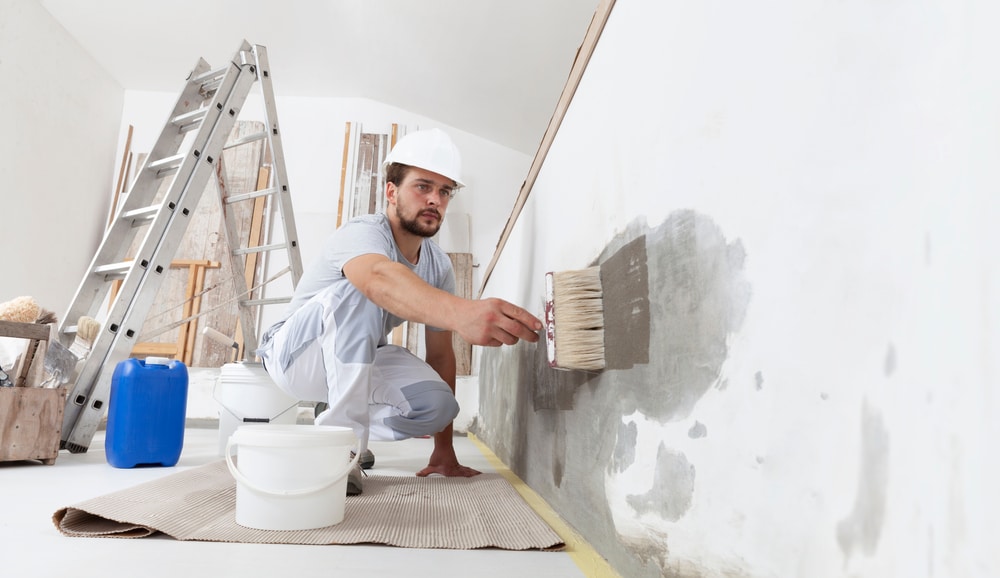 Man applying primer on damaged wall with large brush—illustrates the hidden costs of DIY painting, including time, labor, tools, and surface prep often underestimated by homeowners.

