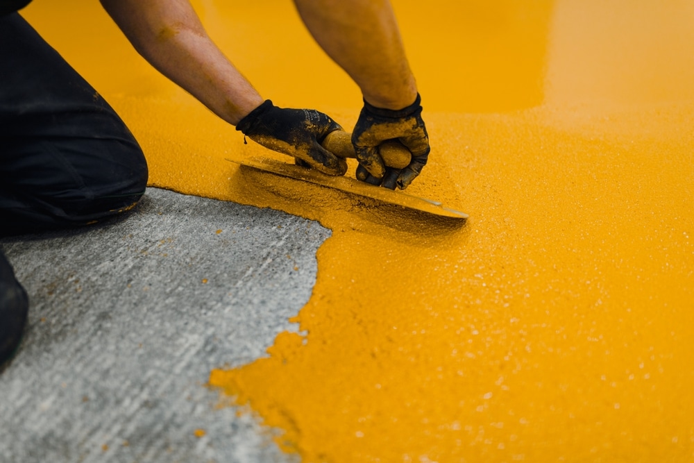 Worker in gloves using a trowel to evenly spread a thick layer of yellow material on a concrete floor, capturing the precision and process involved in professional coating and painting applications for industrial surfaces.

