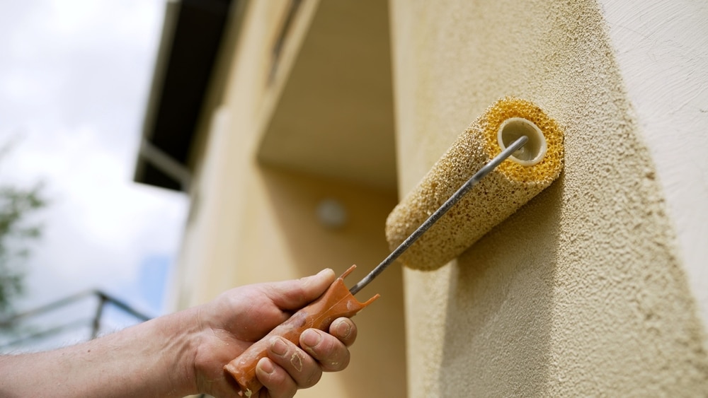 Close-up of a hand applying beige paint with a roller to a textured exterior wall, illustrating a residential painting and coating process for improved surface protection and aesthetic finish.

