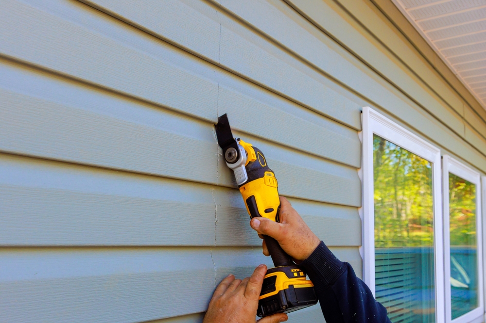 A person uses a power tool to carefully install siding on a house, demonstrating the process of siding installation. The image highlights the precision and effort required to properly fit the panels, ensuring a durable and attractive exterior.







