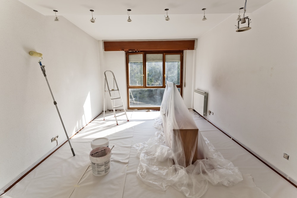 Bright room in the process of being painted white, with plastic sheets covering furniture, paint tools prepared, and natural light streaming through large windows—illustrating the preparation phase common to both residential and commercial painting projects.