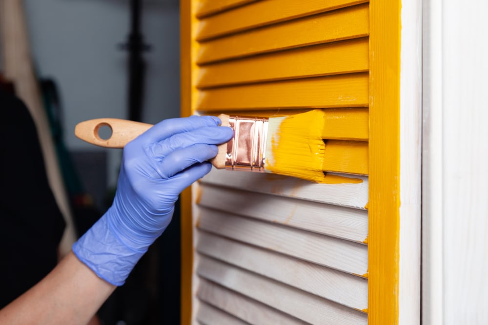 Can I paint over stain without sanding — a person wearing gloves uses a paintbrush to apply yellow paint over a stained wooden shutter without prior sanding.