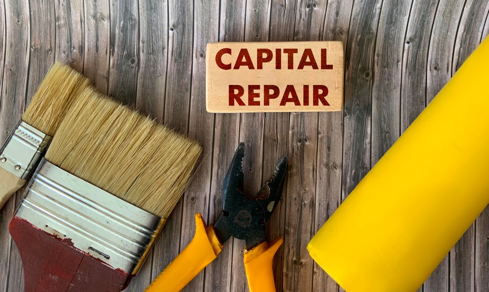 Top-down view of renovation tools including paintbrushes, pliers, and a yellow pipe next to a wooden sign reading 'CAPITAL REPAIR' — representing the question: is painting a commercial building considered a capital improvement