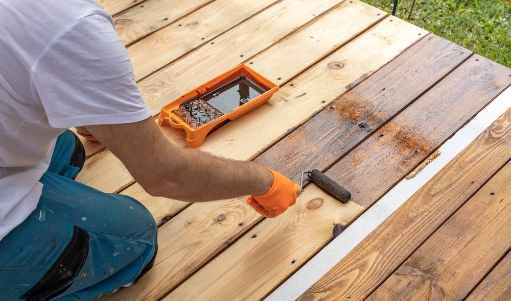 a person kneeling on a wooden deck uses a paint roller to apply stain or sealant, with a tray of product nearby, showcasing the work of professional or DIY deck painters.