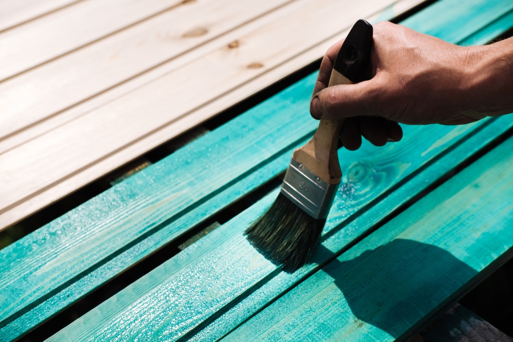Hand brushing vibrant turquoise paint onto wooden planks—step-by-step visual for how to paint a deck.
