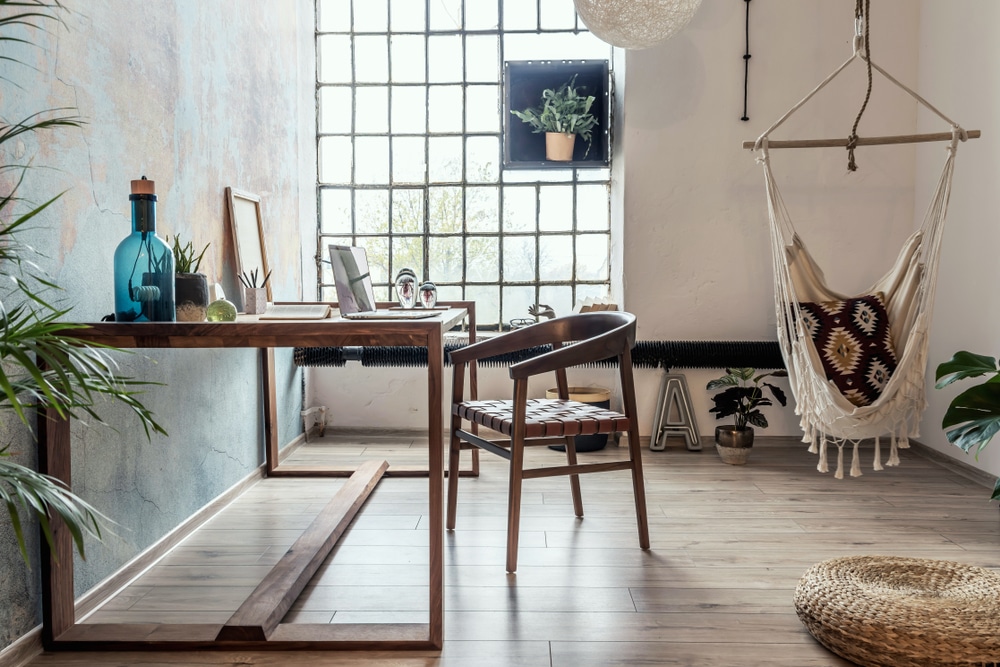 Bright and cozy workspace with natural light, wooden furniture, hanging chair, and indoor plants in a loft-style room — an example of modern home renovations blending comfort and style.