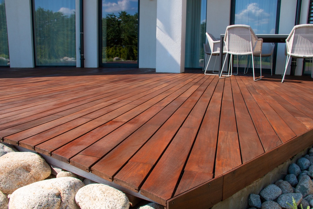 Freshly refinished wooden deck with a rich, dark finish, showing the results of applying stain over paint. The smooth, even tone enhances the outdoor space beside a modern home with patio furniture and large windows.
