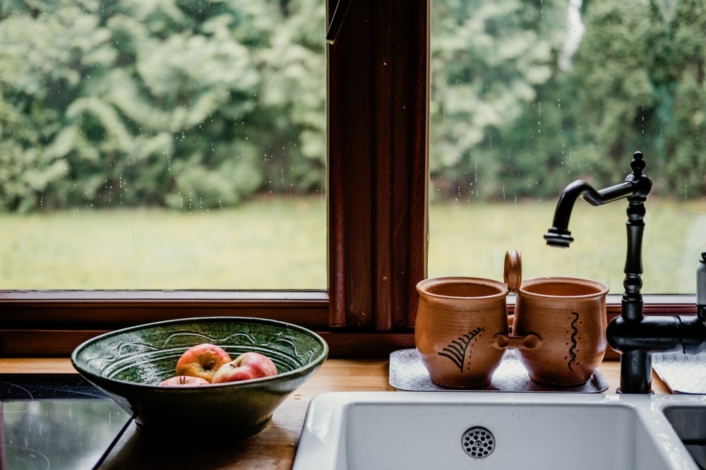 Cozy kitchen sink area with ceramic mugs, a bowl of fresh peaches, and a black modern faucet in front of a large window overlooking greenery—capturing rustic charm blended with modern renovation ideas.