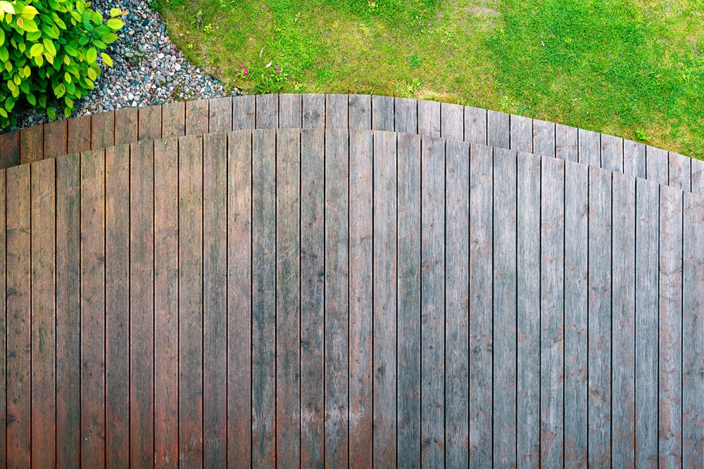 Top view of a curved wooden deck next to green grass and a garden bed, showing an ideal surface to ask how long does deck paint take to dry after application in an outdoor setting.