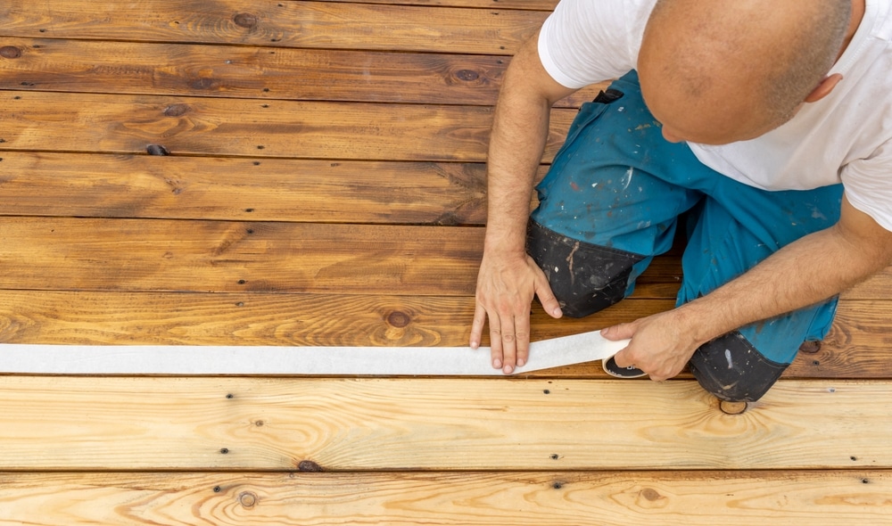 Deck painter kneeling and applying tape to wood boards, prepping for staining or painting.