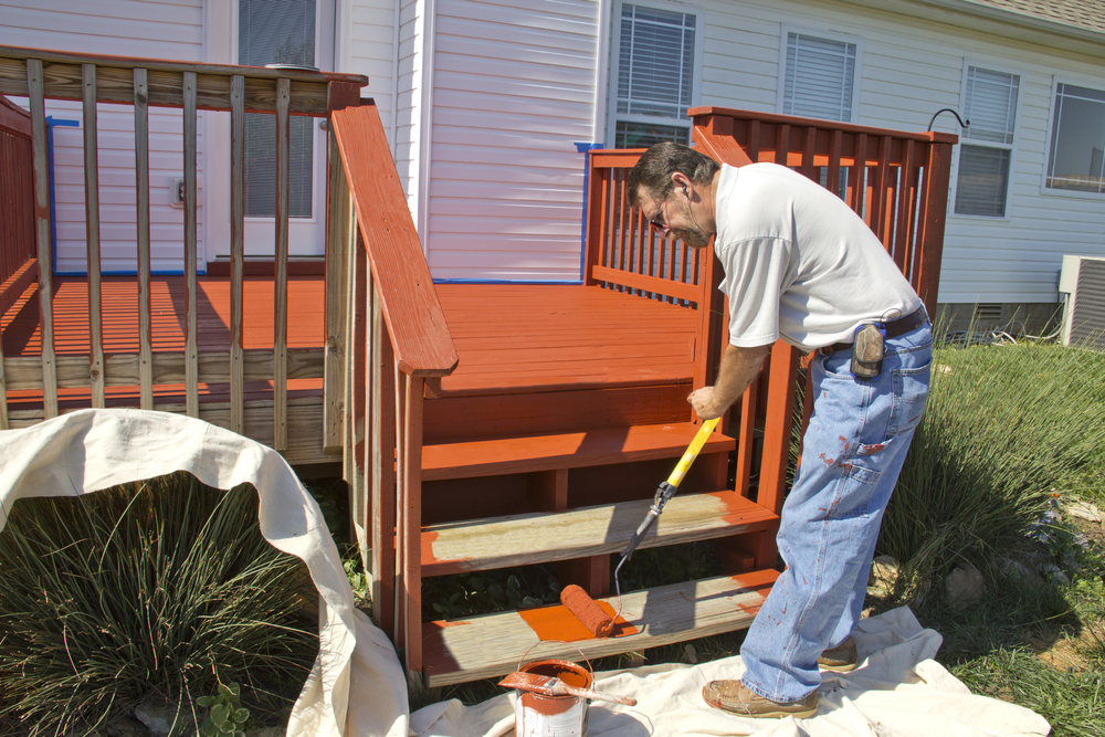 A man staining outdoor wooden deck stairs with a roller brush as part of a home improvement project. This image visually represents the stain vs sealer decision, showing how stain adds rich color and wood protection, while a sealer would preserve the natural look without changing the wood’s tone.