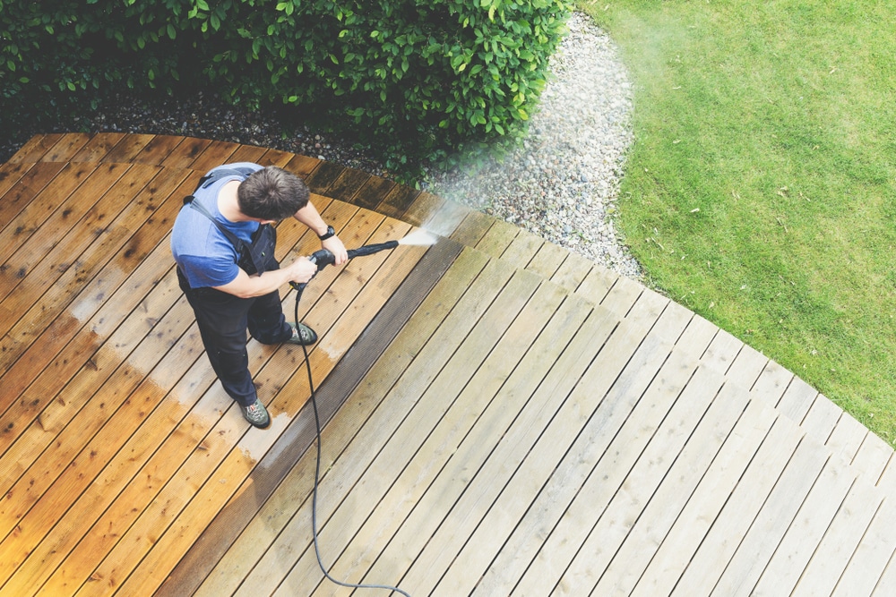 Worker using a high-pressure washer to clean a wooden deck, with a bright clean strip contrasting dirty boards—visual for comparing power washing cost vs. standard pressure washing.