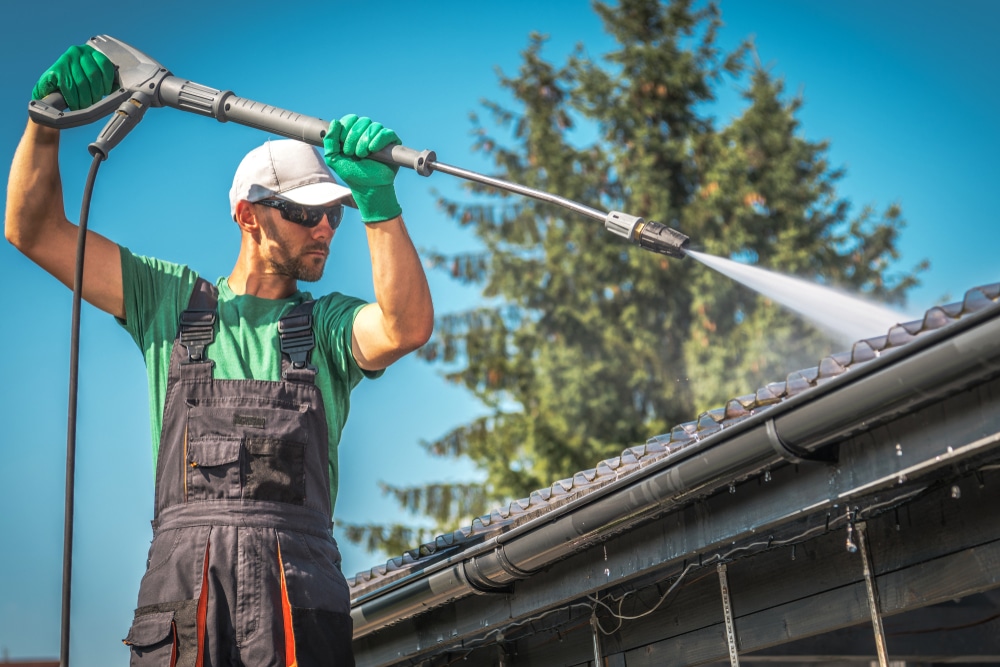 A worker in protective gear uses a high-pressure washer to pressure wash a house roof and gutters, removing dirt and buildup.