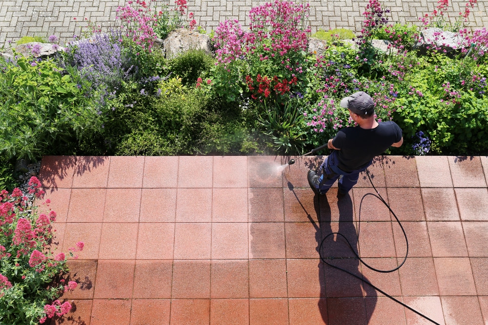 Man using a power washer to clean outdoor patio tiles next to a garden, illustrating how often should you power wash your house to maintain curb appeal and prevent dirt buildup.
