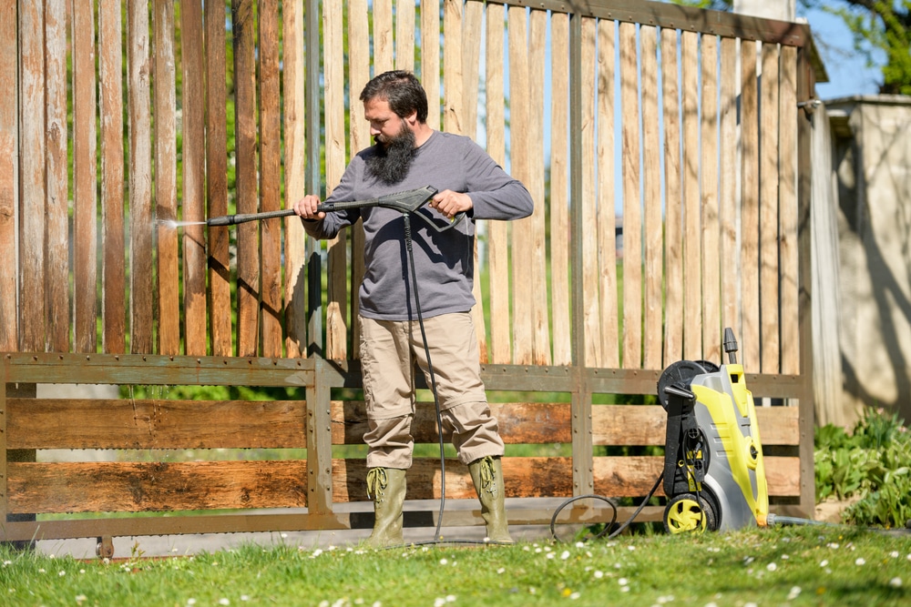 A man uses a pressure washer to clean a wooden fence, demonstrating PSI for pressure washing a house and outdoor surfaces.