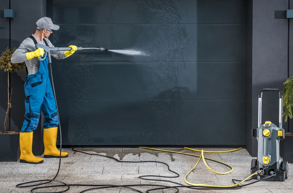 Worker in gloves and yellow boots pressure washing a dark garage door; spray reveals a clean strip, with washer and hoses on the driveway, visual for average cost for pressure washing.
