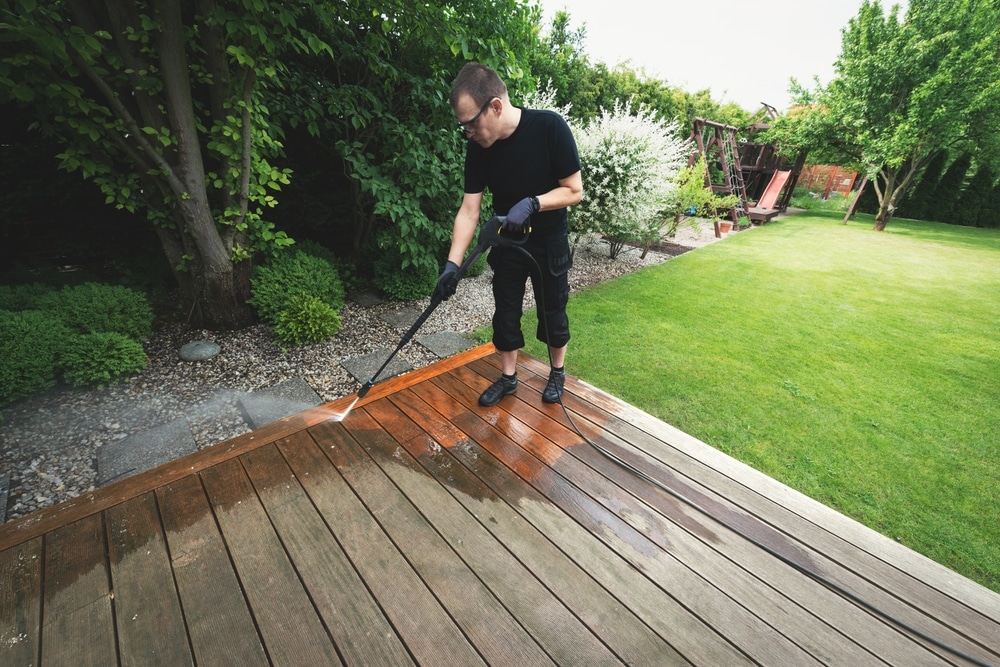 A man uses a pressure washer to clean a wooden deck in a backyard garden, showing the difference in surface color and strength depending on pressure washer PSI.