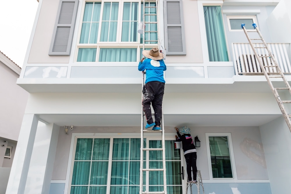 Two workers on ladders painting the exterior of a modern house illustrating a project where homeowners may ask are home improvements tax-deductible.