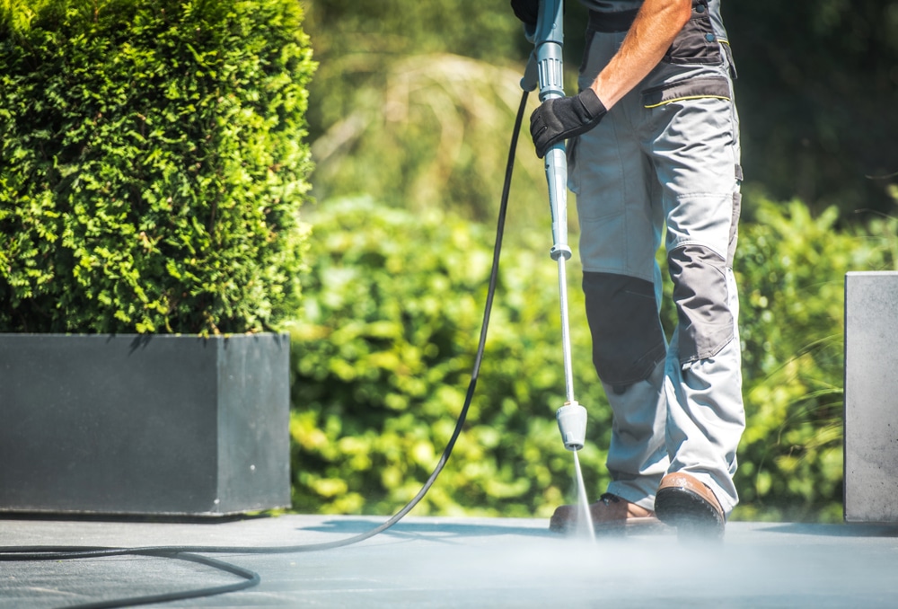 erson using a power washer to clean concrete surface in a garden setting. The question 'Can power washing damage concrete?' can be addressed here, as improper use of power washing equipment, such as using too high a pressure, can lead to surface damage, cracks, or erosion of the concrete.