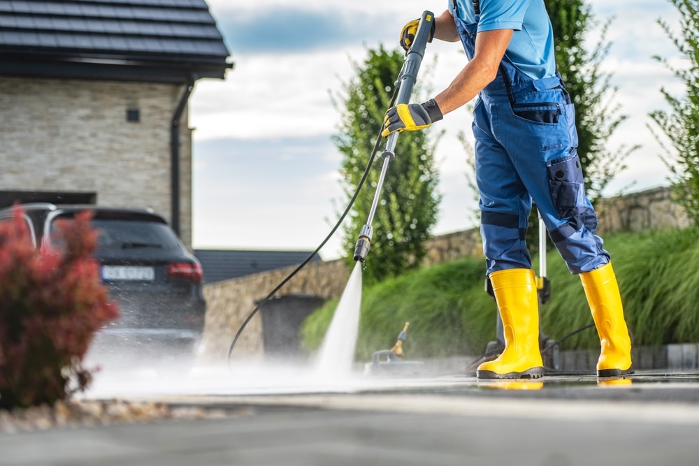 Worker performing driveway pressure washing with a pressure washer, wearing yellow boots and gloves, in front of a car and clean landscape.