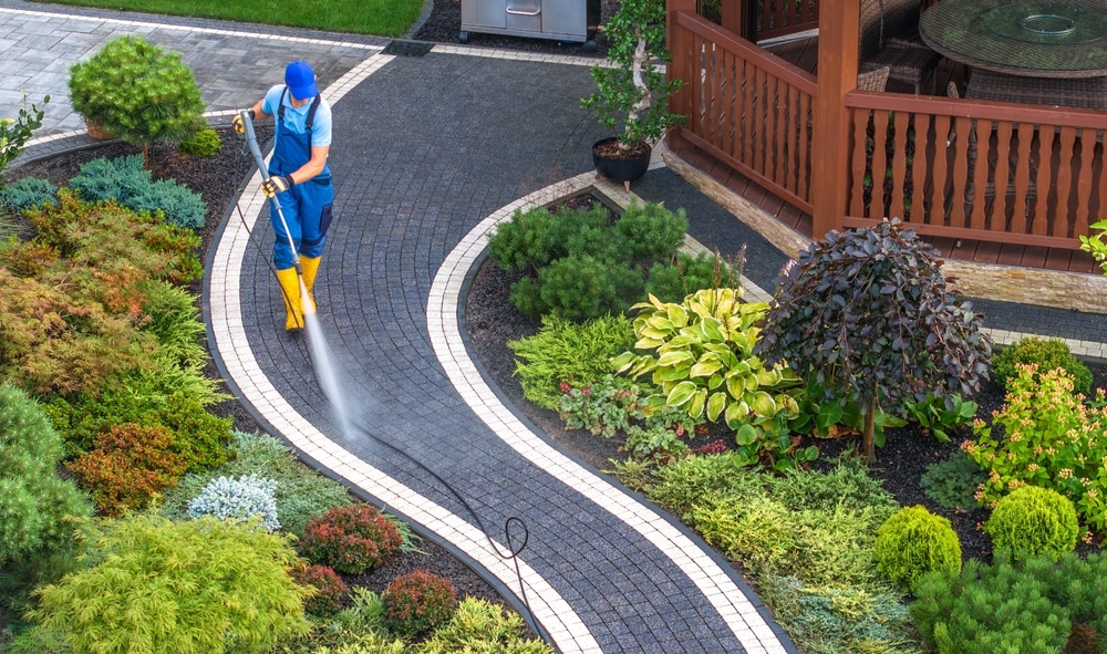 Worker using professional pressure washing equipment to clean a curved stone pathway in a landscaped garden with vibrant plants and shrubs.
