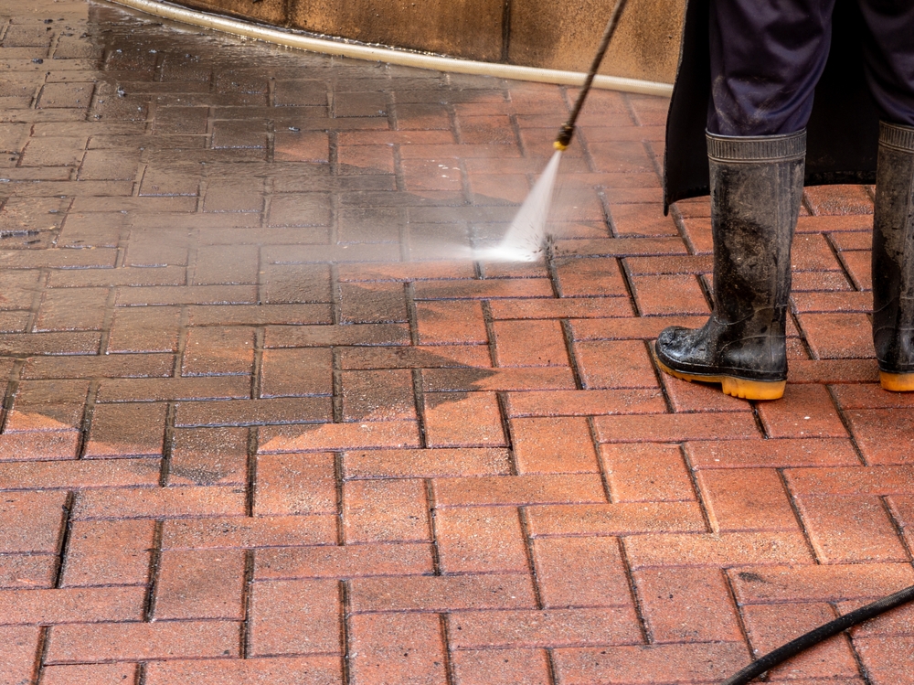Worker using a high-pressure washer to power wash brick pavement, showing a clear contrast between the cleaned and dirty surface.