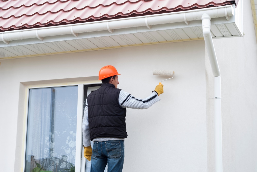 A professional painter wearing a safety helmet and gloves uses a roller brush to paint the exterior wall of a modern house. The image illustrates preparation, technique, and safety—qualities homeowners should look for when researching how to find a painter for reliable exterior painting work.