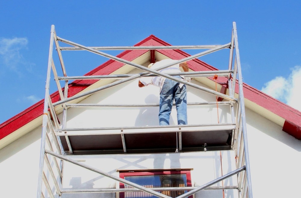 Professional painter standing on scaffolding, painting the exterior of a house with red trim under a clear sky, representing reliable painting companies near me.