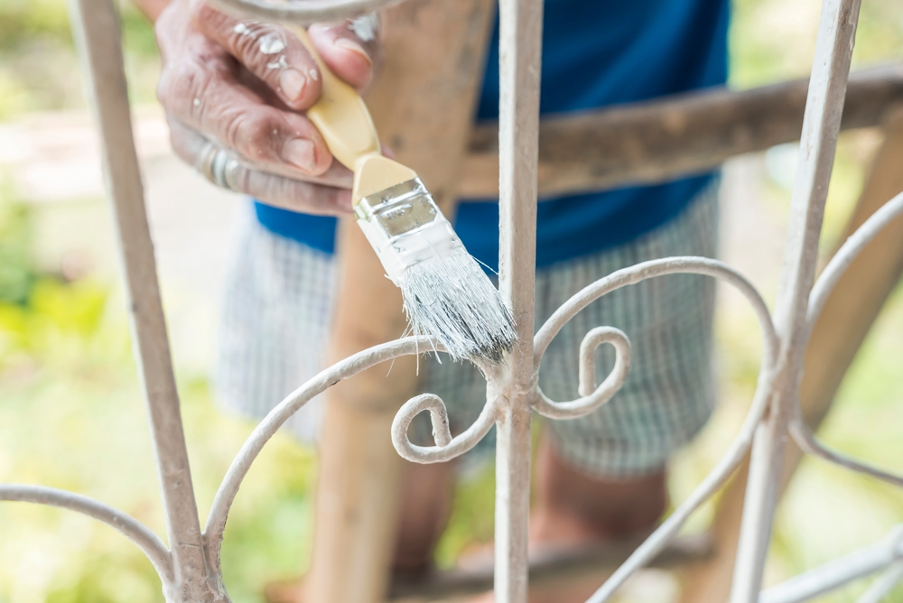 Close-up of a person doing exterior painting, applying white paint with a brush on a decorative metal fence outdoors.
