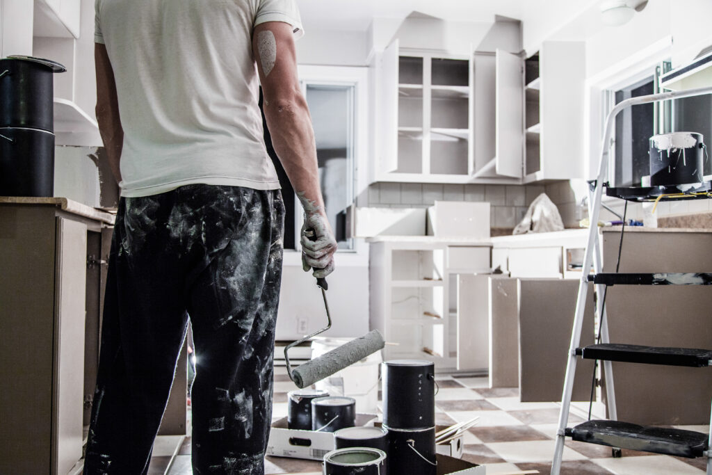 Painter holding a roller and standing in a messy kitchen mid-renovation, surrounded by paint cans and tools — demonstrating pro painting tips for a flawless finish.