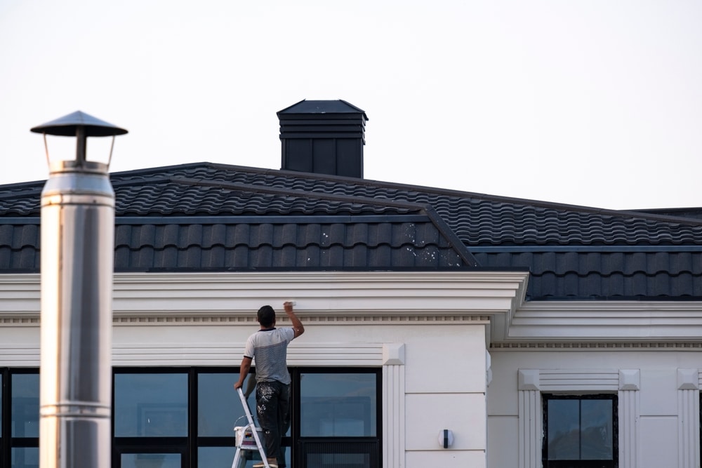 A professional painter stands on a ladder applying paint to the exterior trim of a modern house with a dark tiled roof. The scene captures precision and dedication, illustrating the high-quality workmanship offered by house painters near me who specialize in residential exterior projects.