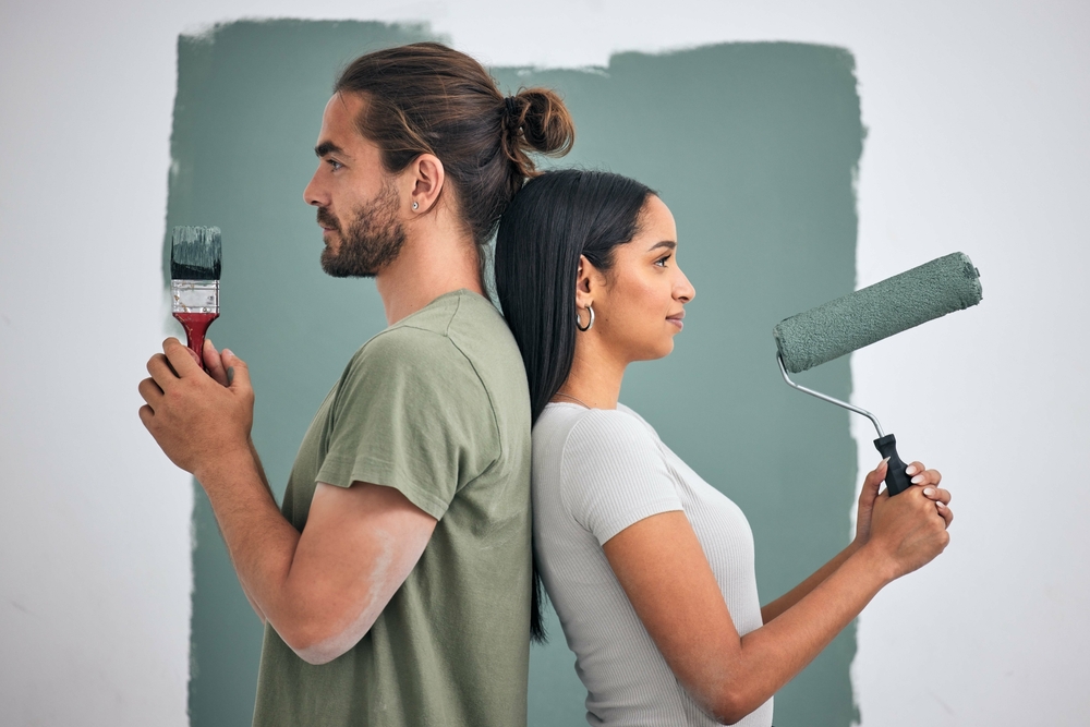 Couple standing back to back holding paint tools in front of a freshly painted wall — demonstrating how to professionally paint a room together with precision and style.