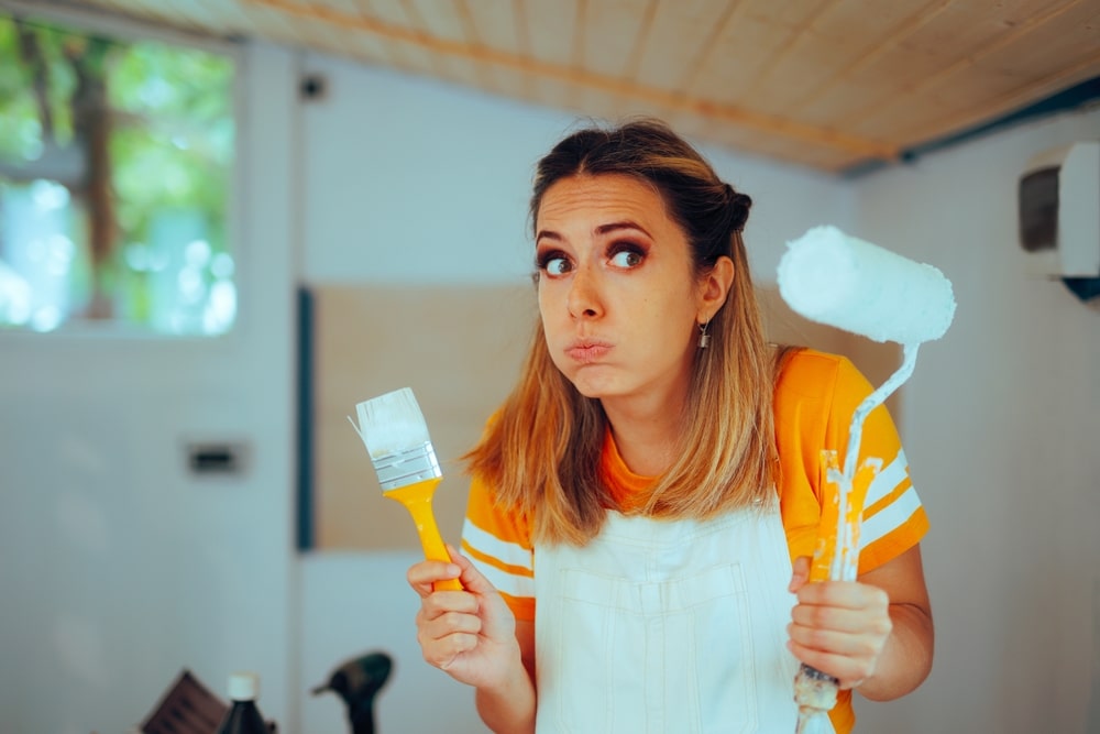 Confused woman holding a paintbrush and roller while painting indoors, illustrating the question 'can you use outdoor paint inside' with a doubtful expression.