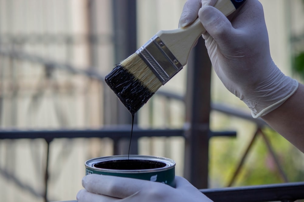 A close-up of a professional painter dipping a brush into a can of black paint while wearing protective gloves. The image highlights precision and care, representing the attention to detail and craftsmanship of reliable painters working on a metal railing project.
