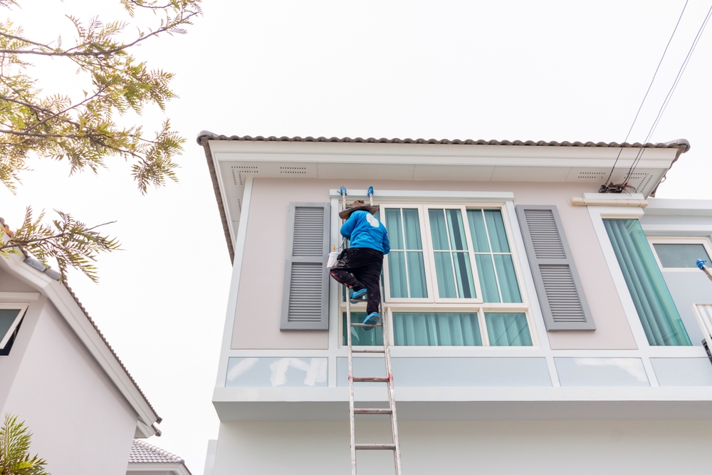 Homeowner on a ladder painting the upper wall of a house, showing how to paint a house exterior yourself safely and efficiently.