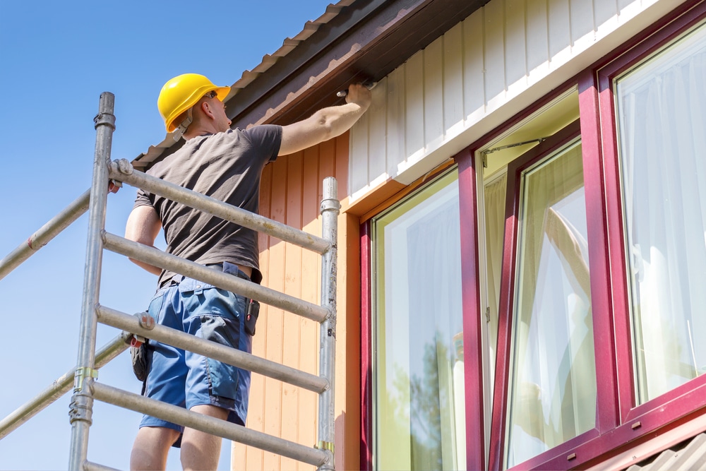 Professional house painter wearing a yellow helmet standing on scaffolding while painting the exterior wall of a modern home, representing skilled house painters at work.