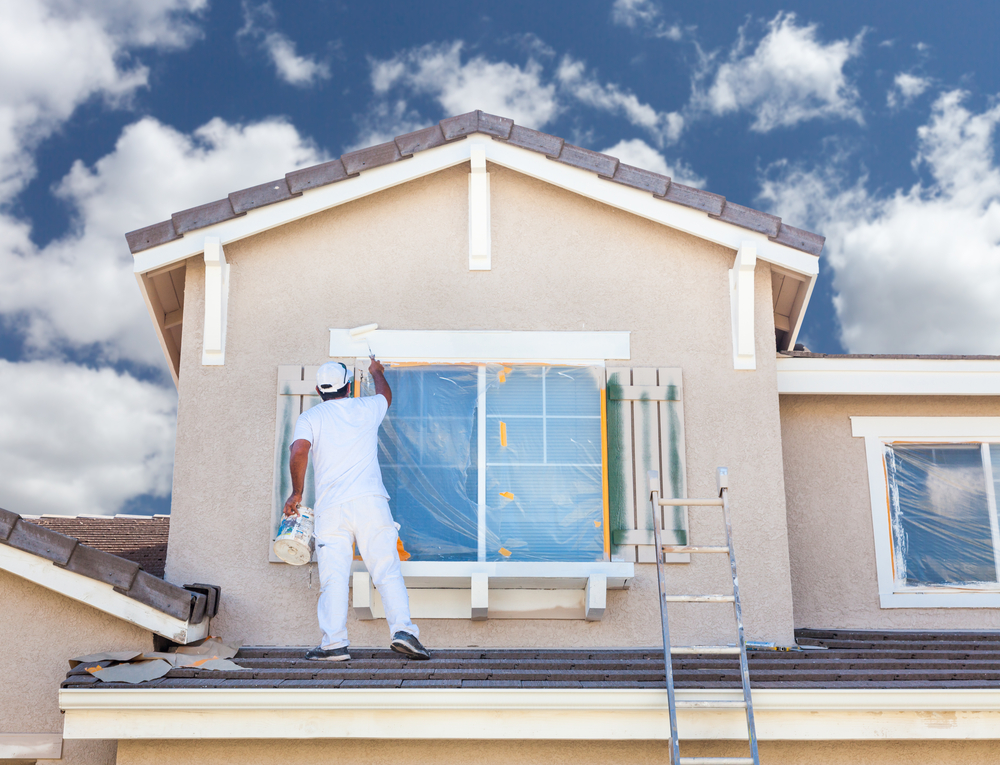 Professional exterior painter standing on a roof and painting the upper wall of a house with a roller, demonstrating expert home painting services.
